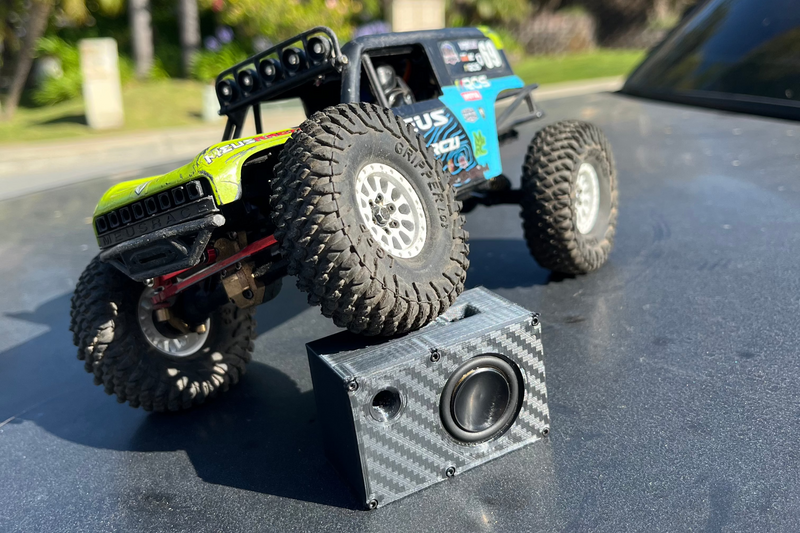 A green and blue comp crawler climbing over The Mini, which sits driver-up on the tarmac. 1/24 and 1/18 scale.
