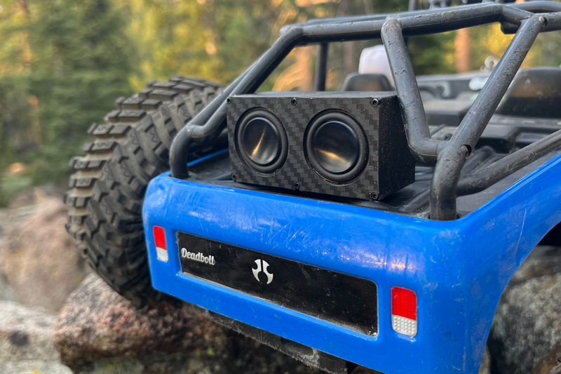 Close-up of The Double sitting in the bed of a blue RC crawler, carbon-look face and both drivers visible. 1/24, 1/18 and 1/10 scale.