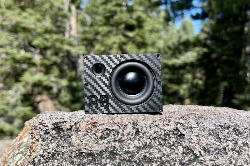 The Super Single on granite with pines behind, one 40 mm driver with a black cone beside a round port. 1/10 scale RC speaker.