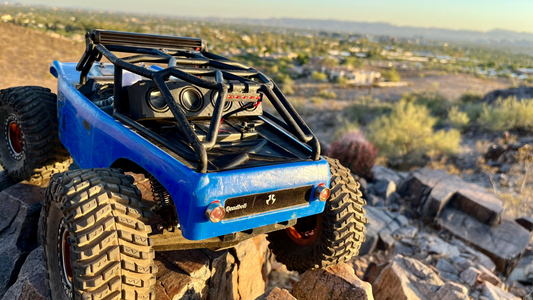 The Quad mounted along the roll cage of a blue RC crawler on rock, valley and city behind. 1/10 scale.