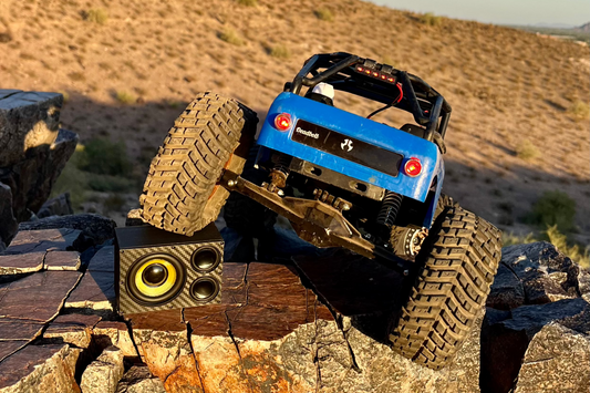 A blue RC crawler climbing broken rock with The Overkill on the slab beneath it. 1/10 scale.