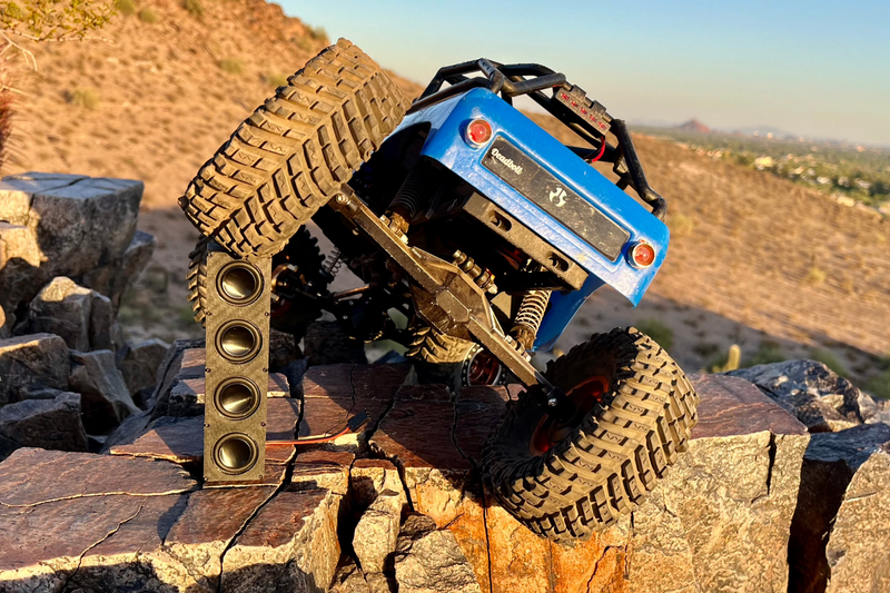 A blue RC crawler climbing a rock face with The Quad mounted across its cage. 1/10 scale.