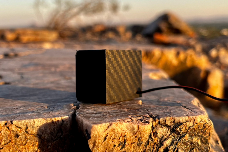 The Micro Single from the side, gold carbon-look weave running across the shell, on a flat desert rock. 1/24 and 1/18 scale.