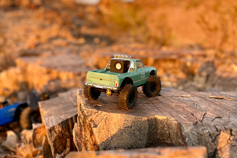 A green Chevrolet-bodied RC pickup crossing broken rock with The Micro Single mounted in the bed, shot from a distance. 1/24 and 1/18 scale.