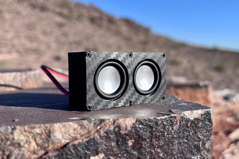 The Micro Double on desert rock under blue sky, two 23 mm drivers side by side in a carbon-look shell. 1/24 and 1/18 scale RC speaker.