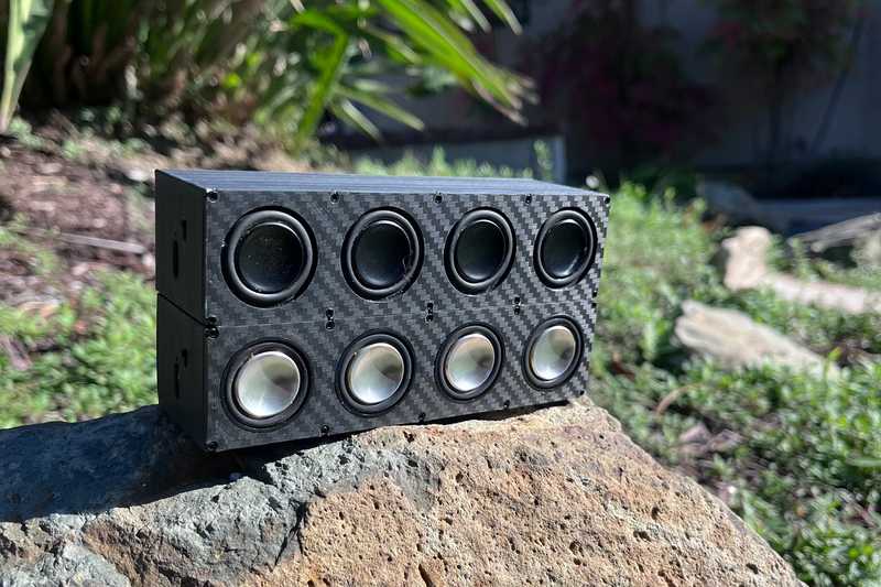 Two Quad speakers propped against a rock in a garden, one behind the other. 1/10 scale.